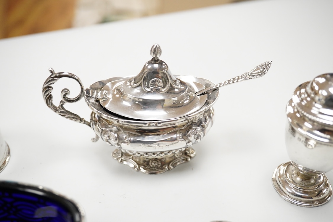 An ornate Edwardian silver oval mustard pot and matching spoon by Barker Brothers, Birmingham, 1902, length, 95mm, together with five other assorted silver condiments including a pair of pierced oval salts with blue glas